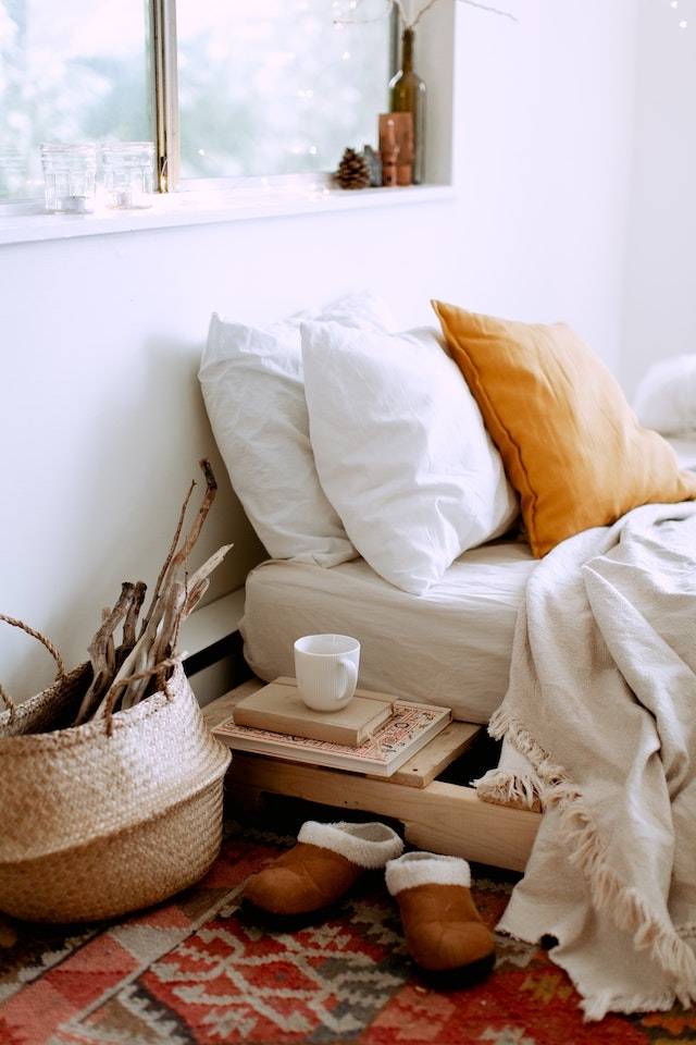 Bed With White Bedding Bedside Table With Books And Wicker Basket And Slippers Next To It